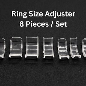 Puede incluir: Un juego de ocho ajustadores de tamaño de anillo de plástico transparente. Los ajustadores son curvos y tienen una pequeña pestaña en cada extremo. El texto "Ring Size Adjuster 8 Pieces / Set" se muestra en blanco.