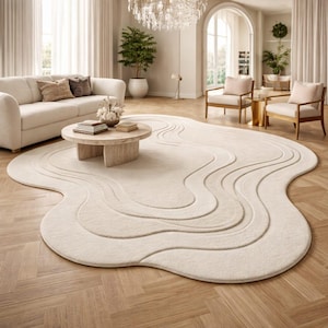 May include: A cream-colored, abstract-shaped area rug with a wavy design. The rug is placed in a living room setting with a sofa, chairs, and a coffee table. The rug's design features multiple layers of raised, textured lines.