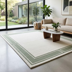 May include: A rectangular area rug featuring a large cream-colored center field surrounded by a sage green border with a geometric frame design. The rug has a low-pile texture and is positioned on a smooth gray floor in a bright living space.
