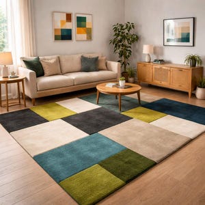 Op de afbeelding: Een moderne woonkamer met een groot vloerkleed met een geometrisch patroon. Het kleed heeft vierkanten in verschillende kleuren, waaronder blauwgroen, groen, grijs en beige. De kamer bevat ook een beige bank, een houten salontafel en kunstwerken.