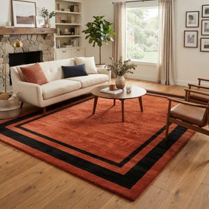 May include: A large rectangular area rug featuring a solid burnt orange centre framed by a thick black border. The rug is placed on a light-toned wooden floor, extending beneath a cream-coloured sofa, a round wooden coffee table, and two chairs.