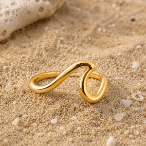 May include: A gold-toned ring featuring a continuous, smooth, undulating wave-like design. The metal surface is polished and reflective, forming a singular, flowing shape that curves upward and downward in a repeating, fluid motion.