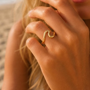 Può includere: Un anello color oro lucido caratterizzato da un design ondulato continuo e liscio che avvolge il dito. Il metallo presenta una finitura riflettente e brillante, creando una silhouette fluida e curva che imita la forma di un'onda oceanica.
