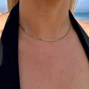 Könnte beinhalten: Eine zarte goldene Halskette. Die Halskette ist im Curb-Chain-Stil gehalten und liegt eng am Hals an. Die Kette hat eine warme Goldfarbe und wird vor einem schwarzen Oberteil und Haut gezeigt.