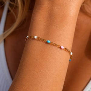 May include: A delicate gold chain bracelet featuring small, evenly spaced enamel beads in white, light blue, and soft pink. The bracelet is worn around the wrist, showcasing a minimalist design with a polished metallic finish and colourful accents.
