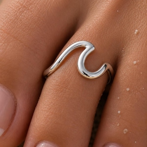 Può includere: Un anello in argento lucido caratterizzato da un design a onda fluido e continuo che avvolge il dito. La superficie metallica è liscia e riflettente, mostrando una forma organica curva che imita la cresta di un'onda oceanica in una fascia.