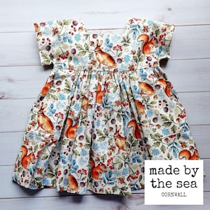 May include: A white dress with a cream background and a woodland print featuring squirrels, rabbits, berries, and leaves. The dress has a square neckline and short sleeves. The dress is made by the sea, Cornwall.