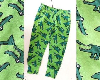 May include: Green children's leggings with a repeating pattern of cartoon crocodiles. The crocodiles are black and white and have open mouths with teeth.
