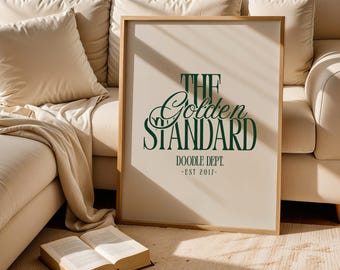 以下が含まれることがあります： 「THE Golden STANDARD」の文字が濃い緑色で書かれたフレーム入りのプリント。下には「DOODLE DEPT. - EST 2017-」と書かれています。プリントは明るい木製のフレームに入っており、ベージュのソファに立てかけられています。開いた本が床に置かれています。