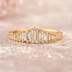 Può includere: Un anello d'oro con una fila centrale di pietre preziose rettangolari e trasparenti, affiancate da pietre più piccole. La fascia è inoltre decorata con piccoli accenti rotondi. L'anello è posto su uno sfondo morbido e scintillante.