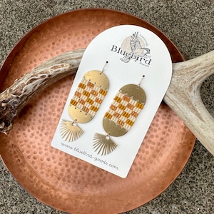 以下が含まれることがあります： 白とオレンジのビーズで幾何学模様が施された、ゴールドのイヤリングのペア。イヤリングは、「handmade Bluebird Goods」と「www.bluebird-goods.com」の文字が書かれた白いカードに吊り下げられています。