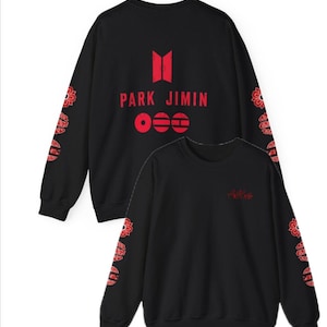Puede incluir: Sudadera negra con texto y gráficos rojos. La parte trasera muestra "PARK JIMIN" y un logotipo. Las mangas tienen un diseño floral rojo. La parte delantera tiene el texto "ARMY" en rojo.