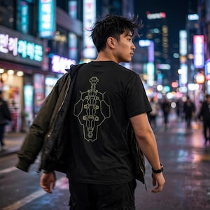 Może przedstawiać: Czarny t-shirt z krótkim rękawem z nadrukiem złotego, geometrycznego, mechanicznego wzoru przypominającego kręgosłup na plecach. Koszulka noszona jest z ciemnooliwkową kurtką zarzuconą na jedno ramię oraz czarnymi spodniami na tle ulicy.