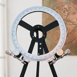 Op de afbeelding: Een navigatie-instrument in vintage-stijl, met een cirkelvormige, gegradueerde wijzerplaat en een zwart spaakontwerp. Het instrument is gemonteerd op een zwarte statiefstandaard. De achtergrond bevat een raam en een kaart.