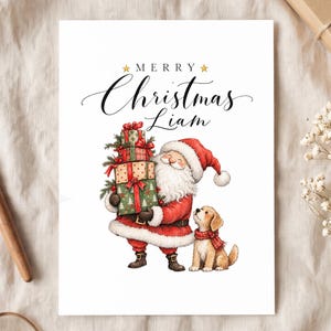 May include: A white rectangular greeting card featuring the text Merry Christmas Liam in black script with gold stars. The illustration shows Santa Claus in a red suit holding a stack of three wrapped gifts next to a small golden puppy.