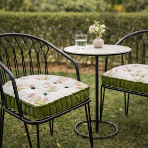 Puede incluir: Dos sillas de metal negro con respaldos arqueados y cojines de asiento capitoné con tela blanca de estampado floral rosa y laterales de terciopelo verde. Una pequeña mesa redonda blanca se sitúa entre las sillas con un vaso y un florero.