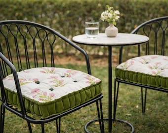 May include: Two black metal chairs with arched backrests and tufted seat cushions featuring a white fabric with a pink floral pattern and green velvet sides. A small round white table sits between the chairs with a glass of water and a vase.