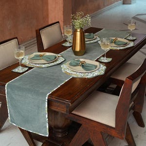 May include: A dining table set for a meal. The table is made of dark wood and has a gray runner. Each place setting includes a plate, napkin, and gold-colored cutlery. Wine glasses and a gold vase with flowers complete the setting.