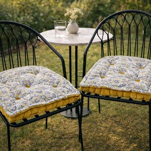 Puede incluir: Dos sillas de metal negro con respaldos arqueados y detalles de alambre vertical. Cada silla tiene un cojín de asiento capitoné con un estampado floral en tonos azul, amarillo y blanco, con un borde de terciopelo amarillo mostaza.