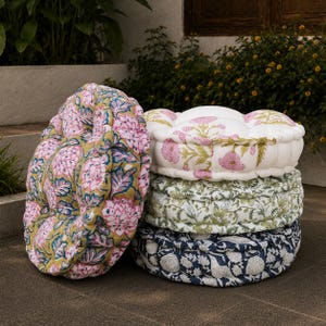 Puede incluir: Una pila de cuatro cojines de suelo redondos con estampados florales de bloque. El cojín superior es blanco con flores rosas, los dos del medio tienen diseños botánicos verdes y blancos, y el inferior es azul marino con motivos florales blancos.