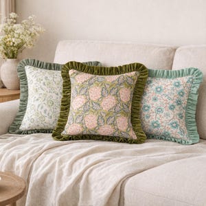 Puede incluir: Tres cojines decorativos con estampados florales y bordes con volantes sobre un sofá crema. Los cojines presentan tonos verdes, rosas y azules, realzando la decoración de la habitación. Los cojines son de forma cuadrada.