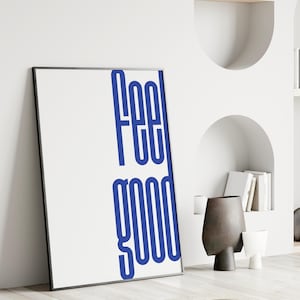Op de afbeelding: Een ingelijste witte kunstprint met de woorden "FEEL GOOD" in vetgedrukte blauwe letters. De print leunt tegen een witte muur, met decoratieve vazen en boeken op planken op de achtergrond. De kamer heeft een minimalistisch ontwerp.