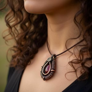 Puede incluir: Un collar con colgante de color cobre con un diseño en forma de lágrima. El colgante presenta una piedra preciosa de color rojo oscuro engastada en un marco de alambre, suspendida de un cordón de cuero marrón. El collar se lleva sobre una parte superior negra.