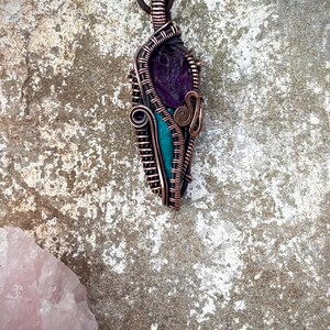 Puede incluir: Colgante hecho a mano con amatista morada y piedra turquesa, envuelto en alambre de cobre. El colgante cuelga de un cordón de cuero marrón. El diseño incluye espirales decorativas y una forma única, mostrando elementos naturales y artesanía.