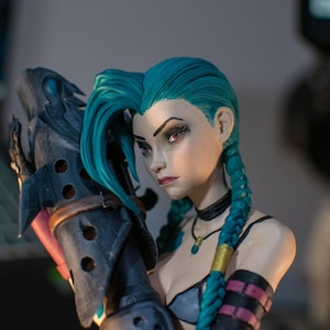 May include: A detailed figurine of a character with vibrant teal hair styled in two long braids. The figure has pale skin, red eyes, and wears a dark choker and necklace. It holds a large, dark, textured weapon with circular cutouts and a brown strap.