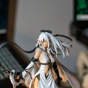 Può includere: Statuetta dettagliata di un personaggio femminile con lunghi capelli bianchi e un abito bianco e nero. La figura ha un fiore tra i capelli e tiene in mano un'arma. Lo sfondo è sfocato, con schermi di computer visibili.