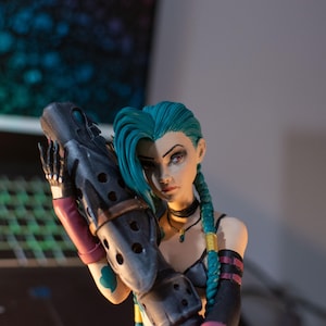Puede incluir: Figura detallada de un personaje con cabello verde azulado en trenzas, sosteniendo un arma grande. La figura tiene una expresión severa y lleva una gargantilla negra y un atuendo oscuro con acentos rosas y dorados. El fondo está borroso.