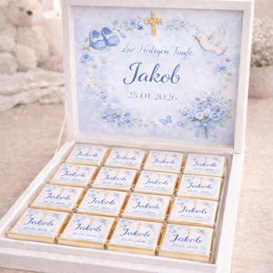 Può includere: Una scatola bianca con un design blu e bianco, contenente piccoli quadratini di cioccolato. Il coperchio della scatola presenta il nome "Jakob" e la data "25.01.2026", insieme a una croce, una colomba e scarpine.