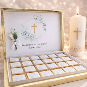Puede incluir: Una caja dorada llena de bombones envueltos individualmente. La tapa presenta una cruz dorada dentro de una corona, con el texto "Kommunion von Ninos 04.05.2024". Una vela encendida con una cruz dorada también está presente.
