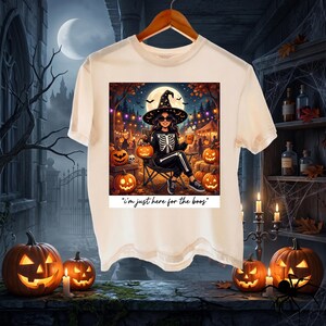 May include: A cream-colored t-shirt featuring a graphic of a person wearing a skeleton-print outfit and a witch hat, sitting in a chair surrounded by pumpkins. The text below the graphic reads, "i'm just here for the boos".
