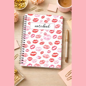 Może przedstawiać: Spiralny notatnik z białą okładką ozdobioną wzorem czerwonych i różowych pocałunków szminki. Słowo "notebook" jest napisane kursywą na białym prostokącie. Złoty długopis i filiżanka kawy znajdują się w pobliżu.
