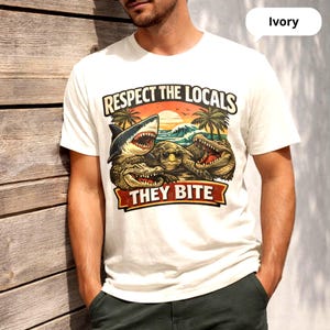 以下が含まれることがあります： サメ、カメ、ワニのグラフィックが特徴のアイボリー色のTシャツ。テキストは「Respect the Locals, They Bite」です。デザインには夕日、ヤシの木、波が含まれています。