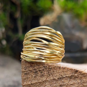 May include: A gold-colored ring with a unique design. The ring is made of multiple thin, wavy bands of metal, creating a textured and layered appearance. The ring is set against a natural wood surface.