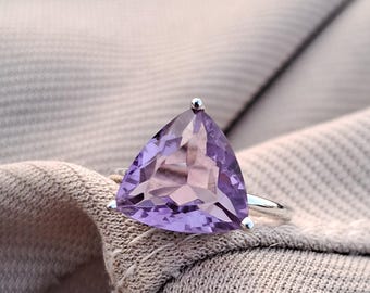 May include: A silver ring featuring a large, triangular, faceted amethyst gemstone. The amethyst is a vibrant shade of purple. The ring is set on a neutral-toned fabric background.