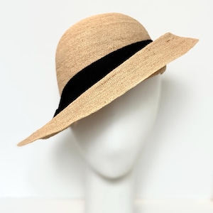 Può includere: Cappello di paglia beige con tesa larga e fascia nera. Il cappello è esposto su una testa di manichino bianca. La tesa è larga circa 10 cm.
