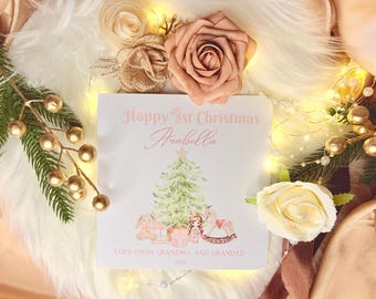 May include: A square white greeting card with pink text reading Happy 1st Christmas Arabella and Love from Grandma and Grandad 2026. The card features a watercolor illustration of a Christmas tree, a plush bunny, a doll, and a rocking horse.