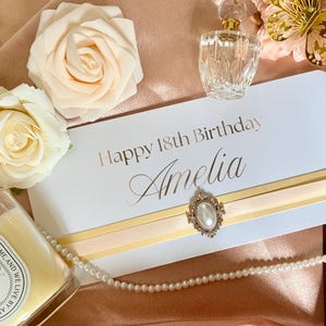 Puede incluir: Una tarjeta rectangular blanca con el texto Happy 18th Birthday Amelia en letras doradas. La tarjeta presenta una banda de cinta dorada horizontal acentuada con un broche ovalado central que contiene una perla blanca. Dos rosas artificiales crema están cerca.