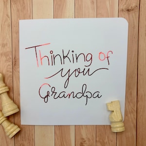 Può includere: Un biglietto d'auguri quadrato bianco con il testo Thinking of you Grandpa scritto in un carattere corsivo rosso metallizzato. Due pezzi degli scacchi in legno chiaro, una regina e un cavallo, sono posizionati vicino agli angoli.
