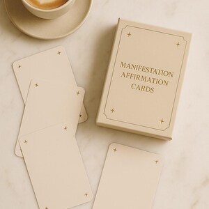 May include: A beige box set of "Manifestation Affirmation Cards" with gold accents, alongside several blank cards with gold star corner details. A cup of coffee with latte art sits on a saucer.