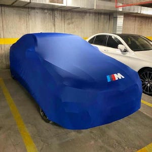 Puede incluir: Una funda azul para coche con el logotipo BMW M en rojo, blanco y azul. La funda cubre un coche en un garaje. Hay un coche blanco aparcado al lado.