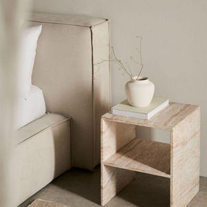 Pode incluir: Uma mesa lateral quadrada de travertino com textura de pedra bege natural e uma prateleira aberta. Em cima, há um livro de tons claros e um pequeno vaso de cerâmica branco-sujo texturizado com um ramo seco fino e delicado.