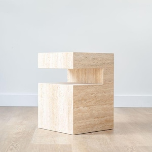 Puede incluir: Una mesa auxiliar de travertino beige con silueta cúbica y un hueco horizontal que forma un estante abierto. La piedra presenta una textura porosa natural con vetas lineales en toda la superficie, creando una forma geométrica sólida y estructurada.