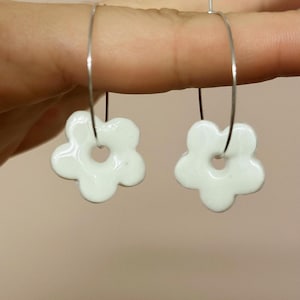 Puede incluir: Un par de pendientes blancos con forma de flor. Cada pendiente presenta un colgante de flor de cinco pétalos blanco brillante, suspendido de un aro plateado. Diseñados como accesorio de moda.