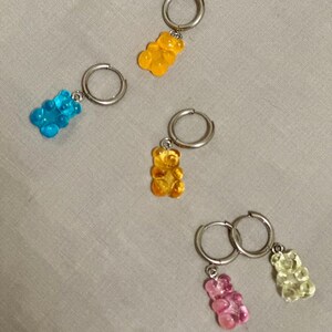 Puede incluir: Un conjunto de pendientes de ositos de goma de colores. Cada pendiente presenta un pequeño dije de osito de goma translúcido en tonos azul, amarillo, naranja, rosa y verde, suspendido de un aro plateado.