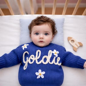 May include: A baby wearing a blue knit sweater with the name "Goldie" in cream-colored script and white daisy accents. A wooden rocket toy rests nearby. The baby is lying on a striped pillow in a crib.