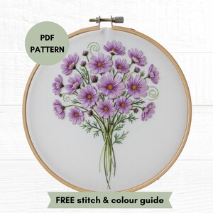 Può includere: Un telaio da ricamo rotondo in legno contenente un tessuto bianco con un bouquet di fiori cosmos viola e steli verdi ricamati a mano. Un badge verde in alto a sinistra riporta PDF PATTERN e un banner verde in basso riporta FREE STITCH & COLOUR GUIDE.